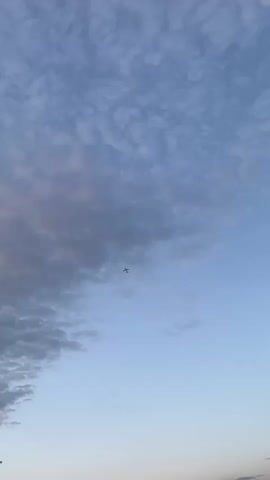 The video shows an airplane flying in the sky. The sky is blue with some clouds. The plane is a small silhouette in the distance. The bottom of the sky is lighter in color, like it is close to sunset.