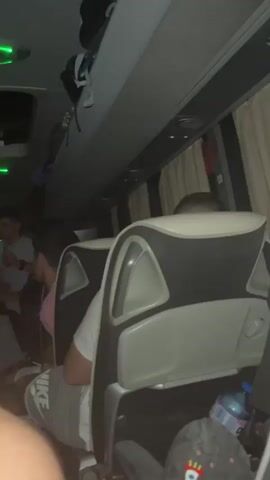 The video shows the interior of a bus at night. Several people are visible sitting and standing in the aisle. The bus has overhead storage compartments and green LED lighting. The seats are white with black accents. The overall lighting is dim.