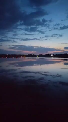 The video presents a serene twilight scene over a body of water. The sky is a blend of dark blues with scattered clouds that reflect faintly in the still water below. The horizon is a soft gradient of pink and orange as the sun sets. Across the water, there's a dark, silhouetted landscape with a few scattered lights that reflect off the water's surface. In the far distance, a faint structure with red lights can be seen. The water reflects the sky and clouds almost perfectly, creating a mirror effect. The foreground is dark, suggesting the viewer is at the water's edge. There are no people or significant moving objects visible in the frame. The overall scene is tranquil and evocative of dusk.