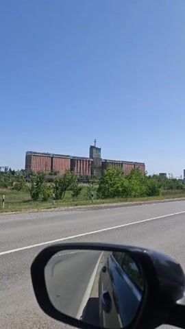 A car drives past a very large grain elevator next to a road. The sky is clear and blue. The elevator is made of brick and concrete and has a tower on top with some sort of antenna. Bushes and small trees are between the road and the elevator. The video is shot from inside the car, and a part of the side mirror is visible. Music is playing.
