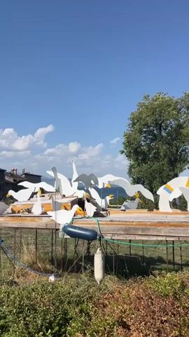 The video shows an outdoor scene on a clear, sunny day with a bright blue sky and scattered white clouds. In the foreground, there is a structure with a wooden platform supported by metal legs. On this platform, there are numerous white, stylized sculptures of seagulls with yellow beaks. These sculptures are arranged in various poses, some appearing to fly or glide. The platform is also adorned with blue and white buoys and green netting. In the background, a church spire with a silver roof is visible to the left, and a large green tree is on the right. Beyond these elements, a body of water and distant hills can be seen. The camera pans slightly across the scene from left to right.