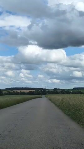 A paved road stretches into the distance, flanked by fields of tall, dry grass under a cloudy sky. In the distance, a treeline with a few structures, possibly houses or farm buildings, is visible. The sky is a mix of blue and white clouds, with some darker, heavier clouds suggesting potential rain. The camera appears to be mounted on a moving vehicle, as indicated by the slight movement and the perspective of the road ahead. No people or other significant objects are immediately apparent in the frame. The overall impression is one of a rural landscape on a day with changing weather.