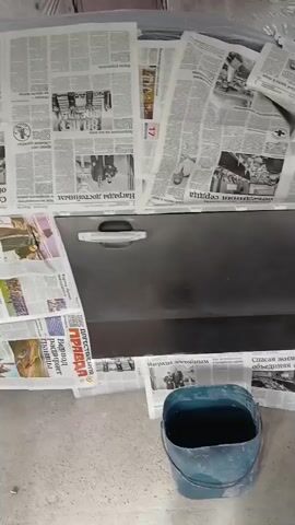 A car door, fender, and hood are covered in newspapers as if for protection or masking. A bucket of dark liquid sits on the floor in front of the car door. The car appears to be undergoing some sort of automotive repair or customization.