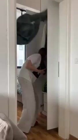 A woman stands in front of an open closet in what appears to be her bedroom. She is wearing a white t-shirt, gray sweatpants, and red slippers. She has long brown hair. The closet is white and contains clothes hanging inside. Behind the woman, through the open doorway, a bed with a patterned bedspread is visible. There is a window in the background, letting in natural light. The walls are white. The woman reaches into the closet, pulls out some clothing, throws it in the air, and states she has nothing to wear. She looks directly at the camera, with her hands on her hips.
