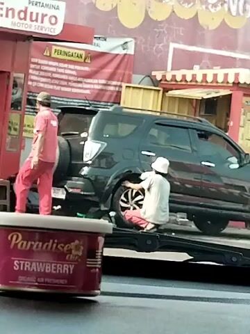 A person wearing a white hat and red pants is kneeling on the ground, working on the front passenger-side tire of a black SUV. Another person wearing a red uniform is standing to the left of the SUV, facing away from the camera and towards the rear of the vehicle. The SUV is parked in front of a building with a red facade and a sign that reads "PERTAMINA ENDURO MOTOR SERVICE". In the foreground, a pink can of "Paradise Air Strawberry Organic Air Freshener" is visible. The overall environment appears to be an outdoor service area or roadside.