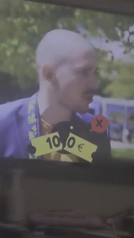 A vertical, full shot shows a man standing in front of a television screen with a video playing. On the screen, a man with a bald head, wearing a yellow shirt and a blue blazer with green and yellow designs, is standing outdoors. He has a mustache and a faint beard. He is facing the right side of the screen. To his right, another man with short dark hair, wearing a black coat, is looking at the bald man. A yellow graphic with black text reading "1000 EURO" and a black graphic with a white "X" are displayed on the screen over the bald man. A yellow banner with black text, "ВЕРЕН ОТГОВОР НАЦИОНАЛЕН ИНСТИТУТ ПО МЕТЕОРОЛОГИЯ И ХИДРОЛОГИЯ (НИМХ)," appears at the bottom of the screen.