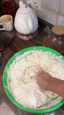 A close-up shot shows a person's hands kneading a large batch of dough in a green plastic bowl. The dough is thick and appears to be made of flour and water, with some parts sticking to the person's hands and fingers. Flour is scattered around the rim of the bowl and on the countertop. In the background, a white teapot, a white mug, and a glass teapot with a wooden lid are visible on a wooden countertop. The kitchen environment appears to be well-lit. The action is focused on the hands and the dough, suggesting a cooking or baking process.