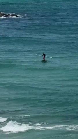 A person is paddleboarding on a choppy teal ocean. The person is wearing red shorts and a dark shirt. They are holding a paddle in their hands and appear to be standing on the board. The water is a dark teal color with visible waves and whitecaps. In the background, there are rocks on the left side of the frame. The video is shot from an elevated perspective, looking down on the ocean.
