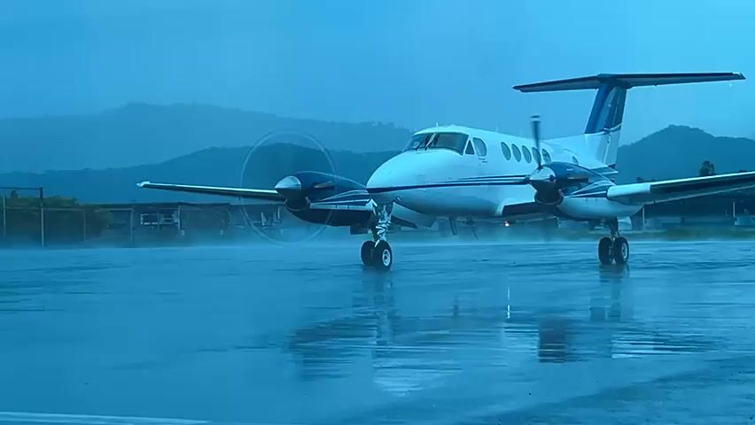 A white and blue twin-engine turboprop aircraft is positioned on a wet asphalt surface under a dark, rainy sky. Its two propellers are actively spinning, kicking up spray from the wet ground. Distant mountains and some structures are visible in the background, partially obscured by the weather. The aircraft appears to be stationary or moving very slowly.