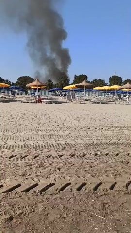 The video shows a beach with rows of lounge chairs and umbrellas under a clear blue sky. In the background, a thick plume of black smoke rises into the air, indicating a fire. The sand in the foreground has tire tracks from a vehicle. The overall scene suggests a sunny day at a beach resort where a fire has broken out nearby.