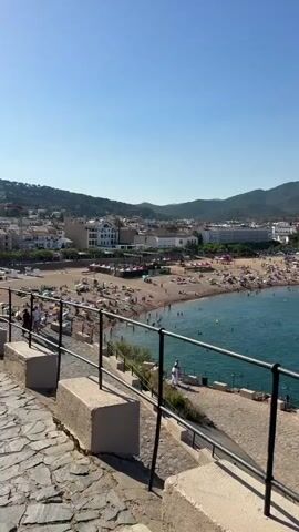 The video shows a panoramic view of a crowded beach and bay on a sunny day. The camera pans across the scene, revealing a coastline dotted with buildings, hills covered in vegetation, and a clear blue sky. The turquoise water is filled with numerous boats of various sizes, anchored or sailing in the bay. The beach itself is packed with people sunbathing, swimming, and relaxing on the sand. In the foreground, a stone pathway and a black metal railing suggest a viewpoint or walkway overlooking the scene. The overall impression is one of a popular tourist destination during peak season, with vibrant activity and beautiful natural scenery.