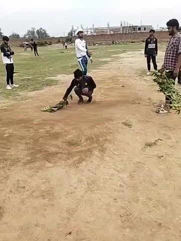 A person is placing leafy branches on the ground. The person is crouched down and appears to be carefully arranging the branches. Other people stand around watching, some holding cricket bats. The scene is taking place on a dirt and grass field with a building under construction in the background.