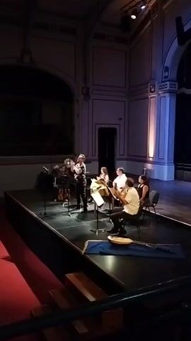 On a stage, a group of musicians rehearses. A man in a black and white floral shirt stands and speaks to the others. He wears glasses and black pants. To his left, a man with long hair sits with a string instrument, and a woman sits next to him. Two men sit opposite, with a woman in a dress seated to the right of them. One of the men plays a string instrument. Music stands are positioned in front of each musician. A long string instrument lies on the floor in front of the stage. The stage is lit by overhead lighting, and there is a large window behind the musicians. The room is ornate with classical architecture.