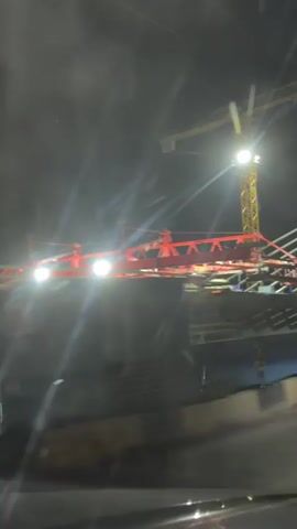 The video captures a nighttime scene of a bridge construction project. A large yellow tower crane is visible, illuminated by spotlights. In the foreground, a red structure, possibly another piece of construction equipment, is also lit up. The bridge itself is partially constructed, with prominent white cables extending from a pylon. The scene appears to be viewed from a moving vehicle, indicated by the motion blur and the angle of the shot. The environment is dark, with the construction site being the primary source of light. There are no visible people or interactions.