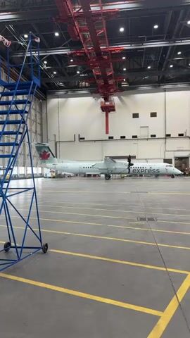 The video displays the interior of an airplane hangar. Two turboprop airplanes, identified by "Canada" and "express" markings, are parked side-by-side on the hangar floor. A large red crane structure is suspended from the ceiling above the aircraft. In the foreground, a blue rolling ladder is positioned to the left. The hangar has a high ceiling with exposed beams and lighting fixtures. The floor is marked with yellow lines, indicating parking or movement zones. The overall environment appears to be an active maintenance or storage facility for aircraft.