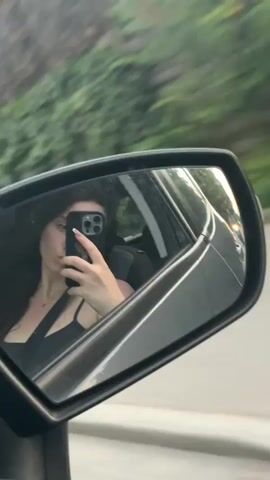 A woman is in the passenger seat of a car, holding a smartphone and taking a selfie in the side mirror. The car is moving, indicated by the motion blur of the trees and buildings outside. The woman is wearing a black top and has long, dark hair. The interior of the car is visible, including the side mirror and part of the dashboard. The scene appears to be during the daytime.