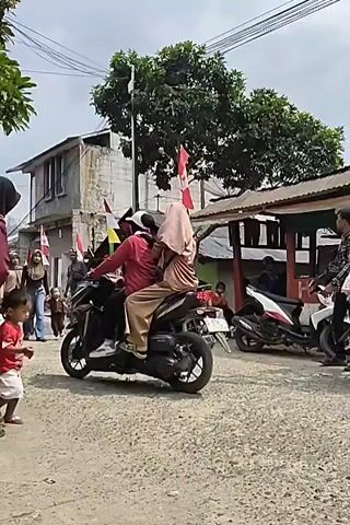 A young child in a red shirt walks towards the camera on a gravel road. Behind the child, a woman in a maroon headscarf and patterned vest walks. Several other people and motorbikes are visible in the background, along with buildings decorated with Indonesian flags.