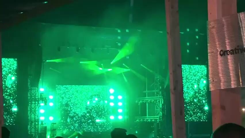 The video shows a concert stage with a DJ performing. The stage is lit with vibrant green, pink, and red lights, with a large screen displaying abstract visuals. The DJ is silhouetted against the bright lights, appearing to be interacting with music equipment. The crowd in front of the stage is dimly lit, with some audience members visible. The overall atmosphere is energetic and typical of a live music event.