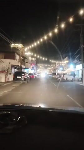 The video is filmed from the perspective of someone in a car driving down a street at night. The street is lined with buildings on both sides, and there are many string lights hanging overhead, creating a festive atmosphere. Several cars are parked along the side of the road and moving down the street. The buildings appear to be shops or businesses, with illuminated signs visible. The overall scene suggests a lively urban environment at night, possibly a commercial district or a street fair.