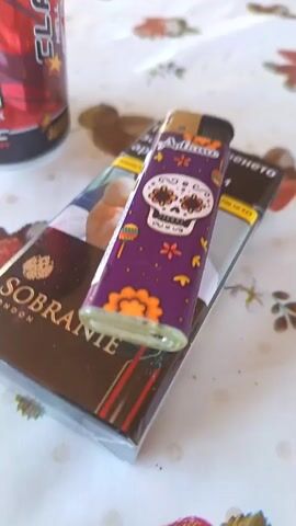 The scene depicts a table setting with several items placed on a patterned tablecloth. The predominant objects are a can of "Hell Energy" drink, a pack of "Sobranie London" cigarettes, and a lighter with a skull design. The Hell Energy drink can is red with black and gold accents, labeled "20 Years Anniversary Classic Energy Drink." The Sobranie cigarette pack is primarily brown, and the purple lighter resting on top features a skull and flower design. The tablecloth has a white base with a pattern of what appears to be strawberries and flowers. No people or actions are visible in the frame.