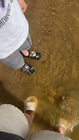 An adult and a child stand in shallow, murky water at what appears to be a lake or river bank. The child, viewed from above, wears a white T-shirt with purple text that reads "ЗАГОРСК," grey cargo shorts, and black Crocs adorned with yellow Jibbitz, one of which says "TEAM." The adult, whose legs and feet are visible in white pants and light-colored sandals, splashes the water with their foot, creating ripples. The adult comments in Russian that the water is "icy" (ледяная), implying it is cold. The ground under the water and on the bank is sandy and speckled with small pebbles. The scene appears to be outdoors during the daytime with bright sunlight.