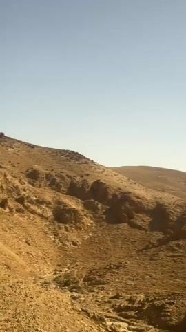 This media file depicts a panoramic view of a dry, desert landscape under a clear, pale blue sky. The scene is dominated by rolling, sandy hills and rocky terrain, with a dry riverbed or gully winding through the lower part of the frame. Sparse, dry vegetation is visible clinging to the slopes. The overall impression is one of a vast, arid expanse.

The activity taking place is not explicitly shown, but the media appears to be a captured moment of observation, possibly a scenic overlook or a view from a moving vehicle. The lighting suggests it is daytime, likely morning or late afternoon given the soft quality of the light and the lack of harsh shadows. There are no people or man-made objects clearly visible in the scene itself, adding to the sense of natural isolation.

The textures of the sand and rock are prominent, emphasizing the ruggedness of the environment. The location cues, such as the arid terrain and the specific geological formations, are consistent with the desert environment of Arad, Israel. The emotions evoked are likely those of solitude, vastness, and the raw beauty of a harsh natural setting. There is no visible text within the media file.