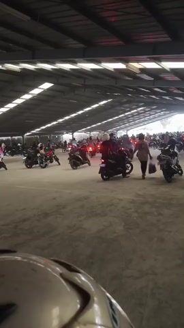 The video shows a large, open-air parking lot or covered area filled with many motorcycles and people. The setting appears to be outdoors under a large, industrial-style roof supported by beams. The ground is paved with a light-colored material, possibly concrete or asphalt. A large number of motorcycles are parked and some are in motion, with riders on them. People are scattered throughout the area, some standing near their motorcycles, others walking around. In the foreground, the helmet of a motorcycle rider is visible, indicating the perspective of someone within the scene. The lighting suggests it is daytime, with sunlight filtering into the covered area from the open sides. The overall impression is of a gathering or event involving a significant number of motorcyclists.