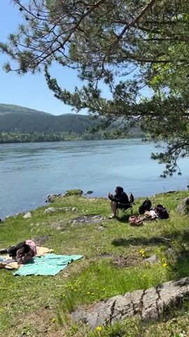 The video shows a serene outdoor scene on a sunny day. Two individuals are relaxing by a wide body of water, likely a lake or river, near Sliznevo, Russia.

One person is seated in a foldable camping chair on the grassy bank, engrossed in their phone. They are wearing a dark hooded top and shorts. Beside them, several backpacks and a brown bag are placed on the ground.

A second person is lying down on a light green blanket spread on the grass, seemingly resting or sunbathing. They are partially covered by a pink or light-colored garment.

The foreground features vibrant green grass dotted with small yellow flowers, and large pine branches with needles partially frame the top of the shot, providing shade. Across the calm, blue water, lush green hills and mountains rise under a clear blue sky, indicating good weather. The overall atmosphere is peaceful and relaxed.