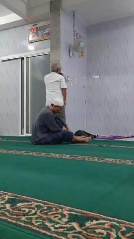 A low-angle medium shot captures two men inside a mosque. The man in the foreground, wearing a grey robe and a white prayer cap, is seated on a green carpet with a patterned border. He is facing away from the camera and appears to be praying. His legs are crossed, and his left hand rests on his knee. A dark bag is placed behind him. The man in the background, dressed in a white shirt and khaki pants, stands facing a wall with his back to the camera. He is looking upwards. The mosque interior features grey tiled walls with a patterned design. A white ceiling fan is mounted on the wall, and a framed clock with Arabic numerals hangs on the wall above a closed sliding door. Several electrical outlets are visible on the wall.
