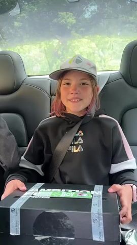 A young girl with pink-streaked hair, wearing a black hoodie and a gray baseball cap, sits in the back seat of a car, holding a black box labeled "Nutrition Boy." She smiles and speaks into the camera. In the background, trees and foliage are visible through the car's windows. Another person's hand holding green binoculars briefly enters the frame on the left side of the screen. The girl adjusts her grip on the box.