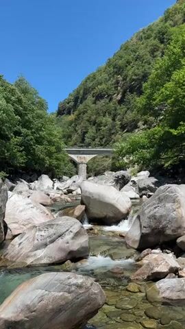 The video shows a clear, rocky river flowing through a gorge. Large, smooth rocks of various sizes are scattered in and around the water. The river is shallow in parts, with visible gravel and smaller stones on the riverbed. Lush green trees line the steep slopes of the gorge, meeting a clear blue sky. A concrete bridge with two visible pillars spans the gorge in the background, with a hint of graffiti on the underside of the bridge. The sun is shining brightly, indicating a clear day. There are no people or animals visible in the frame.
