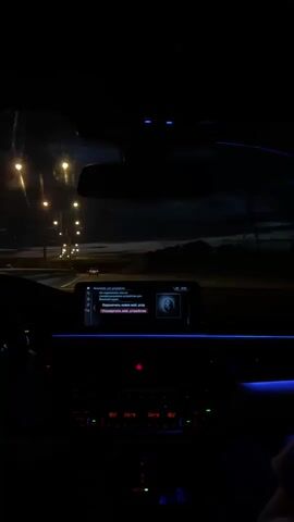 The video shows the interior of a car at night. The driver's hands are on the steering wheel, and the dashboard lights are on, casting a blue glow. The car is moving, as evidenced by the passing streetlights and the slight blur of the scenery outside. A digital display on the dashboard shows a message in Russian, which appears to be an alert or notification. The overall atmosphere is dark and moody, suggesting a nighttime drive.