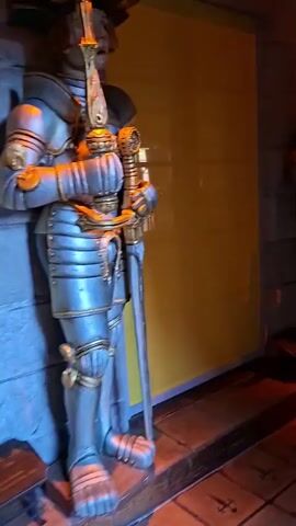 A video shows a hallway lined with life-sized statues of knights in armor. The knights are standing on raised platforms in front of a yellow wall. The hallway has stone walls and a tiled floor with a medieval-inspired pattern. There are also some doors visible at the end of the hallway. The lighting is dim, with spotlights illuminating the statues and creating shadows. The video pans across the hallway, showing the different statues and the overall atmosphere of the space.