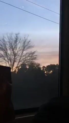 The video shows a view from inside a moving vehicle, likely a train, looking out of a window. Outside, trees and vegetation are silhouetted against a sky with colors of sunset or sunrise, displaying light orange and pale blue hues. The view is somewhat obscured by the glass. Parts of seats are visible inside the vehicle, including a seatback that is orange. Overhead power lines are also visible through the window.