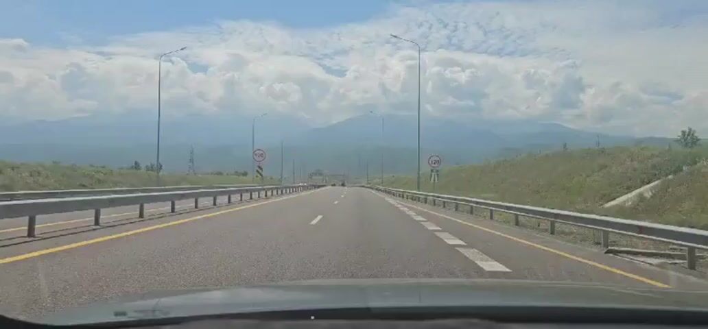 The video captures a first-person perspective of driving on a multi-lane highway. The road is asphalt with clear white lane markings and a yellow line separating lanes. Guardrails line both sides of the highway. In the distance, mountains are visible under a sky with scattered clouds. There are streetlights and utility poles along the road. Speed limit signs indicating 120 are visible on both sides of the highway. A few vehicles are present on the road, and the scenery suggests a rural or semi-rural environment. The overall impression is a smooth, forward motion along a well-maintained highway with natural scenery in the background.