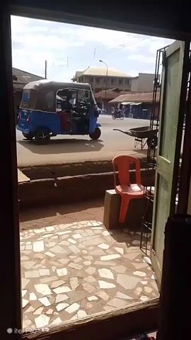 The video shows a street scene viewed from an open doorway. In the foreground, the tiled floor of what appears to be a shop is visible, with an orange plastic chair placed on a concrete block to the right. Beyond the doorway, a dirt road with a few market stalls and buildings lines the street. Several vehicles are present, including a blue auto-rickshaw and a motorcycle. The sky is partly cloudy.