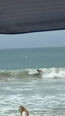 The video shows a wide shot of a beach with the ocean in the background. In the foreground, partially obscured by an umbrella, are three people sitting or kneeling on the sand. The ocean is choppy with white waves. A person on a surfboard is riding a wave. The sky is overcast. The camera is static.