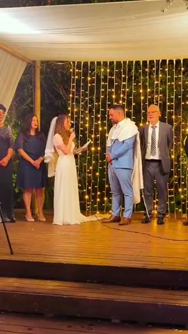A wedding ceremony is taking place outdoors under a canopy decorated with string lights. A bride in a white dress and veil stands at a microphone, reading from a paper. A groom in a blue suit and a tallit stands opposite her. Two women, presumably family members, stand to the bride's left, and a man, likely a family member or officiant, stands to the groom's right. A videographer, wearing a black shirt and grey shorts, moves across the foreground of the scene, filming the ceremony. The ground is a wooden platform.