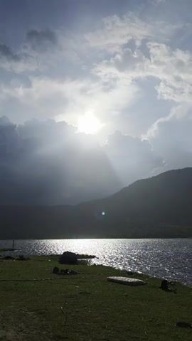 The video shows a lake scene on a partly cloudy day.  The sun is shining brightly, partially obscured by clouds, casting a glare on the water's surface. In the distance, a silhouetted mountain range can be seen.  The foreground features a grassy shoreline. The camera pans upwards to focus on the sun and clouds.