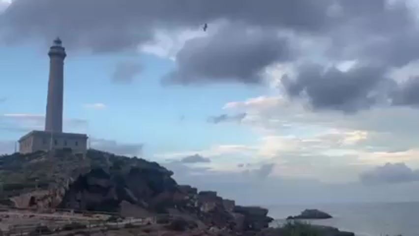 A tall lighthouse stands atop a rocky cliff, overlooking the ocean. A bird flies across the sky in front of the lighthouse. The sky is partly cloudy with hues of blue. The ocean is visible in the background.