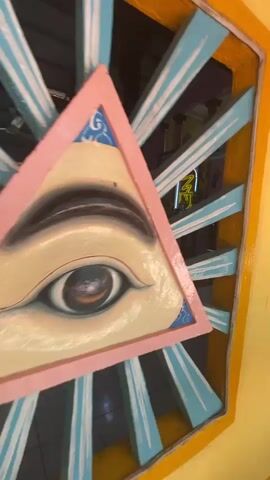 The video pans from a pink framed painting of an eye, through a blue and yellow painted structure, revealing the interior of a building, which appears to be a place of worship or gathering. The building has a tiled floor, pink walls, and a high ceiling with visible wooden beams. There are blue-draped tables, fans, and decorative flags hanging from the ceiling. In the center of the room, there's a raised platform or stage with a glowing yellow sign. The interior architecture includes arched doorways leading to other rooms.