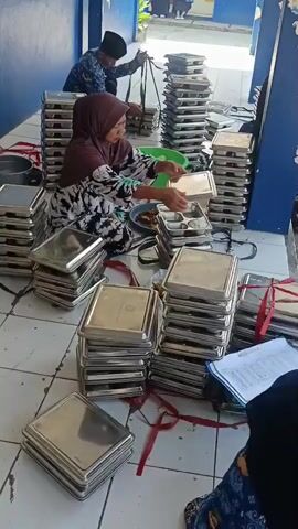The video captures an outdoor scene where several stacks of metal trays are arranged on the ground. In the foreground, a woman wearing a headscarf and a patterned dress is seated on the ground, sorting through items placed on a tray. To her left, a large green bowl contains white rice, and next to it are several small containers with food. In the background, a person in a uniform is interacting with stacks of trays, appearing to be organizing or moving them. Another individual, also in uniform, is visible further in the background. The environment appears to be an outdoor walkway or courtyard with a blue wall on the left and a yellow building structure in the distance. Red ribbons are visible tied around some of the stacks of trays. A person's arm and shoulder are visible in the bottom right corner of the frame, holding a document. The overall impression is of a food preparation or distribution activity involving a large number of trays.
