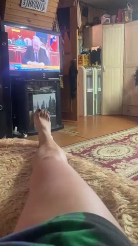 The video shows a person's legs stretched out on a fuzzy rug, with a television screen in the background displaying a news broadcast. The person appears to be relaxing or watching TV. The room has a rustic feel, with wooden paneling on the ceiling and walls. There are several objects visible, including a TV stand with a picture frame, cabinets, and a doorway. The lighting suggests it could be daytime or evening. The TV screen shows a man in a suit, likely a politician, speaking at a podium. Text on the wall above the TV appears to be in Russian, possibly a decorative sign with a phrase about home. The overall atmosphere is casual and domestic.