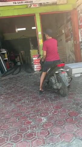 A person is sitting on a black motorcycle outside a store. The person is wearing a maroon t-shirt and black shorts. The store has a green awning and is filled with various items, possibly building materials. The ground is paved with hexagonal red tiles. The motorcycle's brake lights are on.