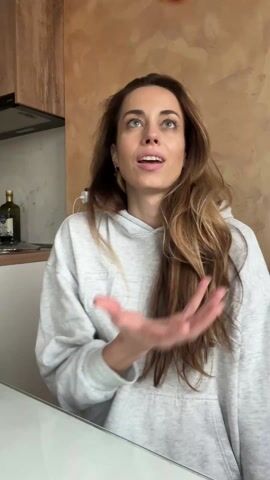 The video shows a woman seated at a white table holding a yellow and red apple, which she takes a bite out of. She is wearing a light gray hooded sweatshirt. She has long brown hair and is facing the camera. In the background, there is a kitchen with a counter, a stovetop, and a bottle of olive oil. The wall behind her has a textured, tan finish.