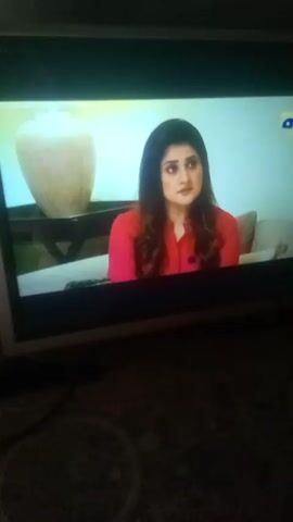 The video shows a television screen displaying scenes from a drama or soap opera. The scenes shift between three different women. The first woman is wearing a bright pink shirt and sits on a couch next to a decorative vase. The second woman is an older woman wearing a white shirt and glasses. The third woman wears a light green dress with a floral print and raises her hands while speaking emphatically. The background of the scenes varies but appears to be interiors of homes or studios. The television screen has a dark bezel. The video is shot indoors with poor lighting.