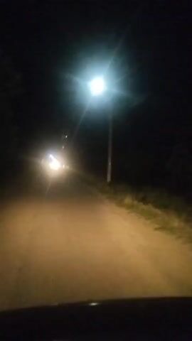 The video captures a nighttime drive on a rural road near Tamarisi, Georgia. The scene is dominated by the darkness of night, with a single streetlamp illuminating a portion of the road and its surroundings. The camera perspective suggests it is filmed from inside a vehicle, likely from the dashboard, as the lower part of the frame shows the car's interior.

The primary focus of the video is the road ahead, which is lit by the vehicle's headlights, creating a warm, hazy glow. In the distance, the headlights of another vehicle are visible, creating a bright, star-like effect due to the camera's exposure. The road appears to be unpaved, with vegetation and tall grass lining the right side. The overall atmosphere is quiet and solitary, typical of a rural setting at night. There is no visible text in the media file. The time of day is clearly night, and there are no indications of specific weather conditions, though the haziness could suggest a slight mist or dust in the air. The emotion conveyed is one of a peaceful, perhaps even slightly eerie, journey through a dimly lit landscape.