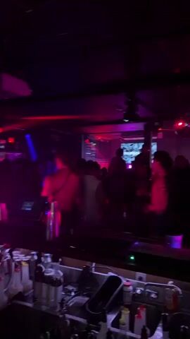 The video shows a crowded nightclub with a DJ booth in the foreground. The DJ, a man with his back to the camera, appears to be shirtless. He is interacting with equipment in front of him. The club is lit with purple and pink lights, and a disco ball is visible in the background, reflecting light. Several people are dancing in the dimly lit space. The overall atmosphere is energetic and typical of a nightclub setting.