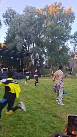 The video shows an outdoor event with a stage in the background. Several people are present, some dancing or performing on the grass in front of the stage. One person is wearing a beige jumpsuit, another a black top and leather pants, and two others are in bright athletic wear. A person in a blue outfit is on the ground. Two smartphones are being held up in the foreground, presumably recording the event. The environment appears to be a park or open grassy area with trees and buildings visible. The lighting suggests it is late afternoon or early evening.