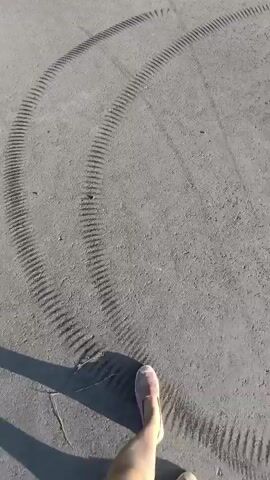 The video captures a first-person perspective of someone walking on an asphalt road. The road is bordered by lush green vegetation and trees that form a canopy overhead, creating a dappled light effect on the ground. Tire tracks are visible on the road surface, suggesting recent vehicular activity. The camera angle pans slightly, giving a sense of movement and the progression of the walk. The lighting indicates it is daytime, likely late afternoon or early morning due to the angle of the shadows.